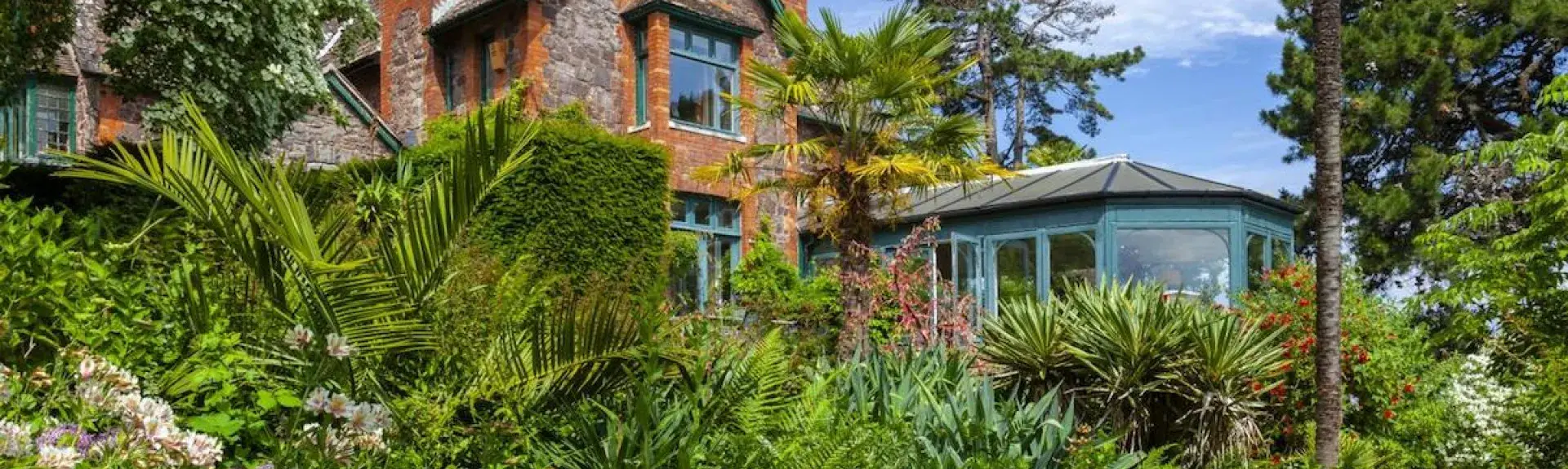 A House with large conservatory is surrounded by a garden with lawns and mature shrubs.