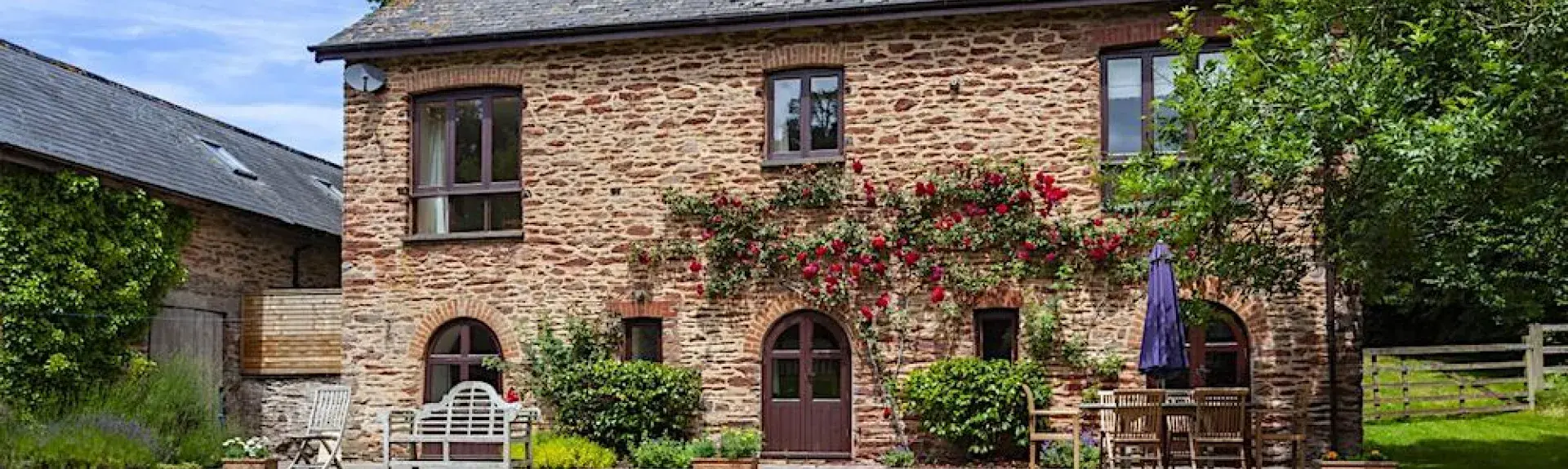 A  stone-built cottage overlooks a large lawn surrounded by flowering shrubs and bushes.