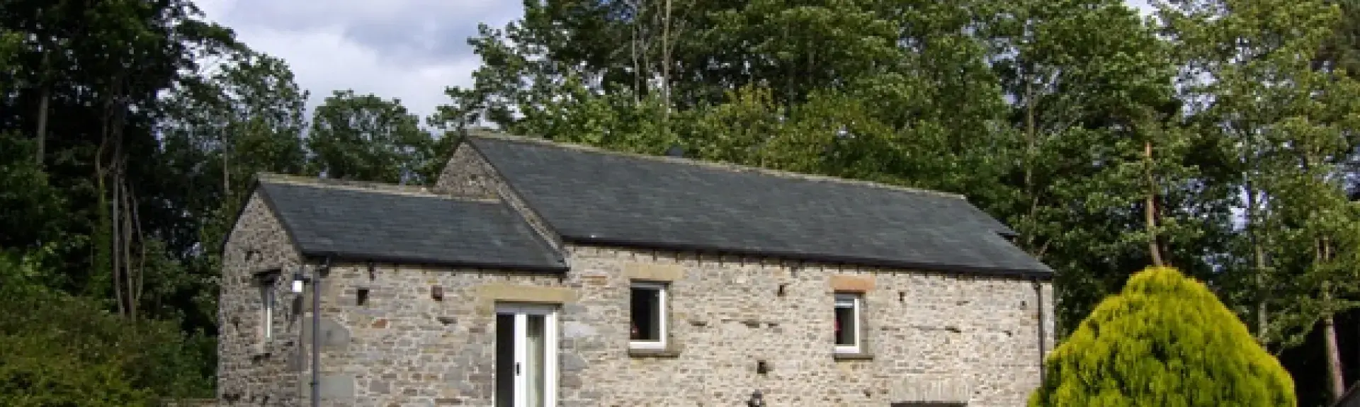 Doiuble-fronted exterior of a stone holiday cottage in Cumbria surrounded by mature trees.