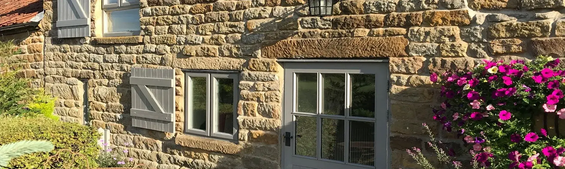 Exterior of a sandstone cottage with a courtyard lined by flowering shrubs.