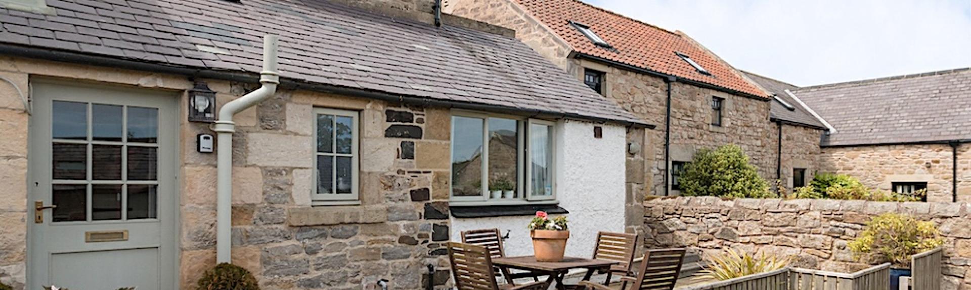 Exterior of a stone-built holiday cottage with ground fllor extension behind a paveed  garden with dining table and chairs.