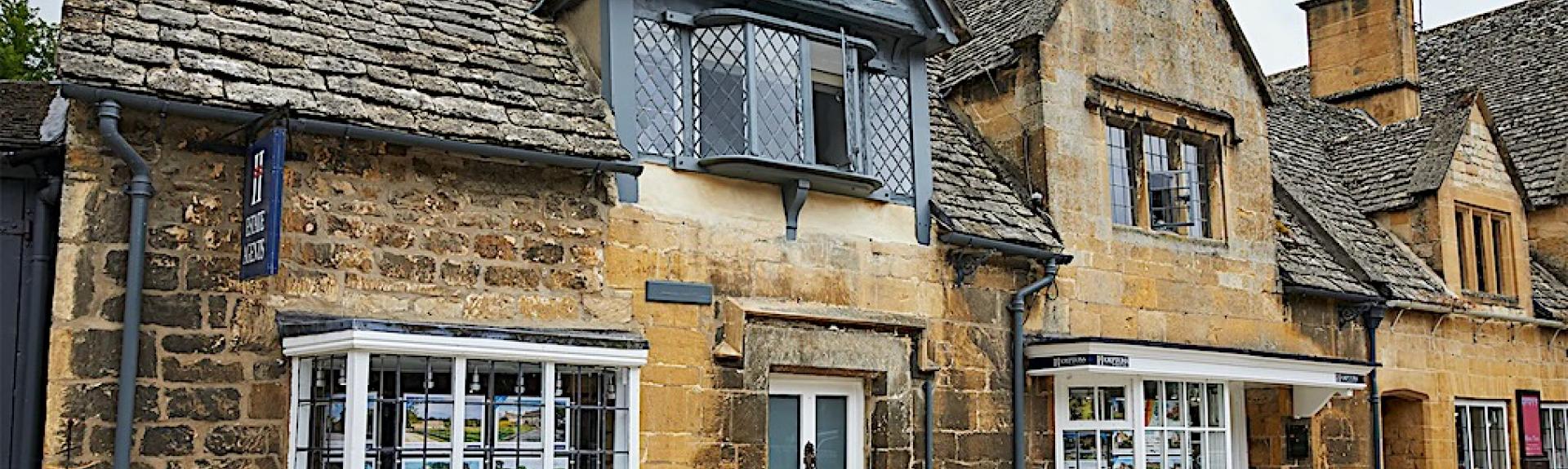 A semi-detached Cotswold holiday cottage with leaded, bay windows overlooks a village street.