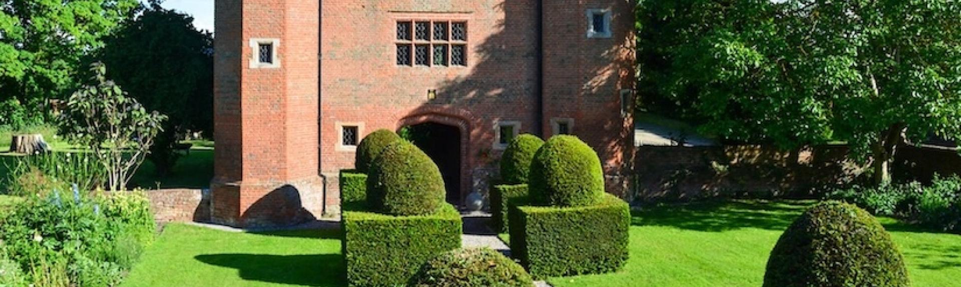 A grand 3-storey, brick-built Tudor house overlooks a large, formal lawned gardens.