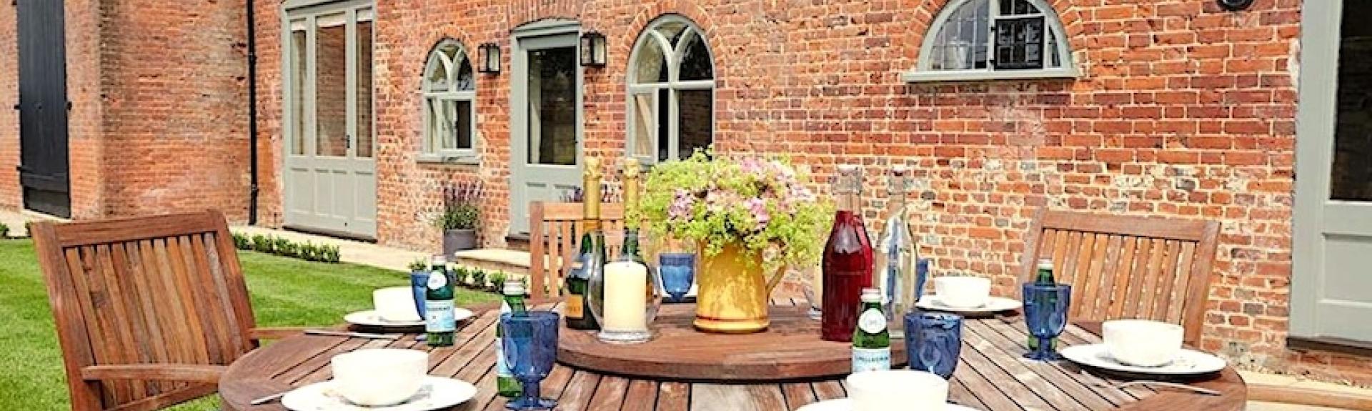 A wood dining table set for breakfast stands on a courtyard in front of a brick-built Norfolk cottage with arched windows.