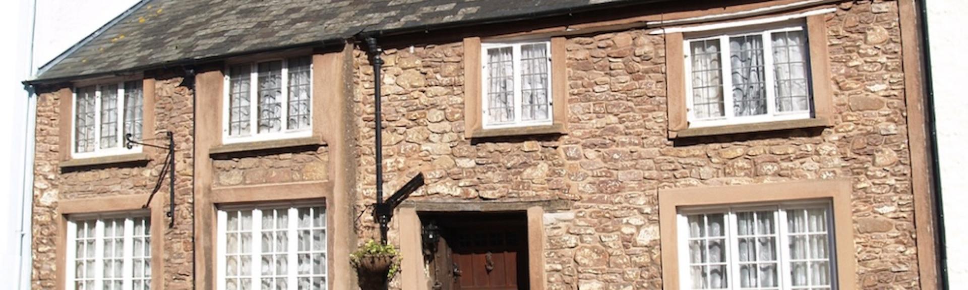 Exterior of a double-fronted stone holiday cottage in Dunster nestling into a steep wooded hillside.