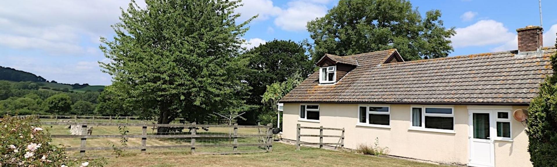 A single-storey Dorset holiday cottage overlooks a large lawn surrounded by a wooden fence, trees and rose bushes.
