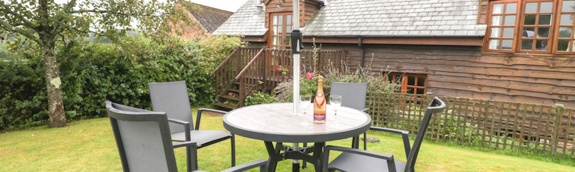 A wooden holiday cottage overlooks a large lawn with outdoor dining furniture.