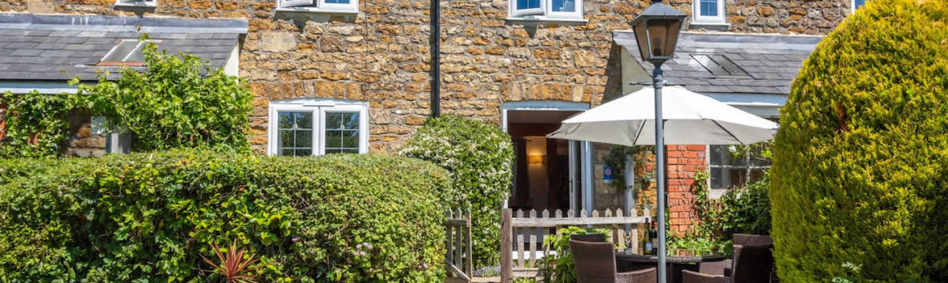 Exterior and front garden of a stone-built Dorset holiday cottage with outdoor dining furniture on its terrace.