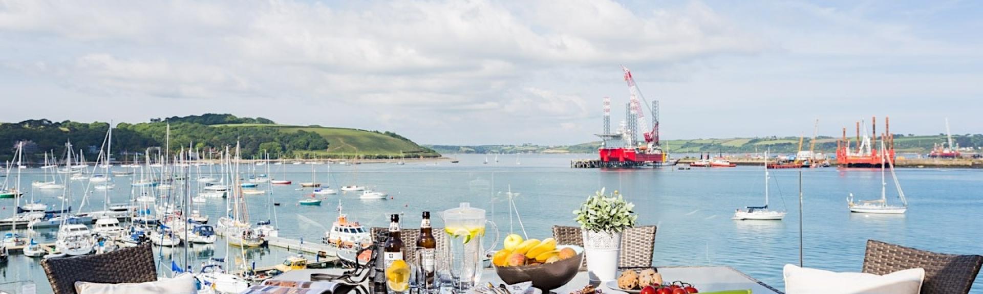 An outdoor table laid for dinner on a deck overlooks a boat-filled Falmouth esturary.