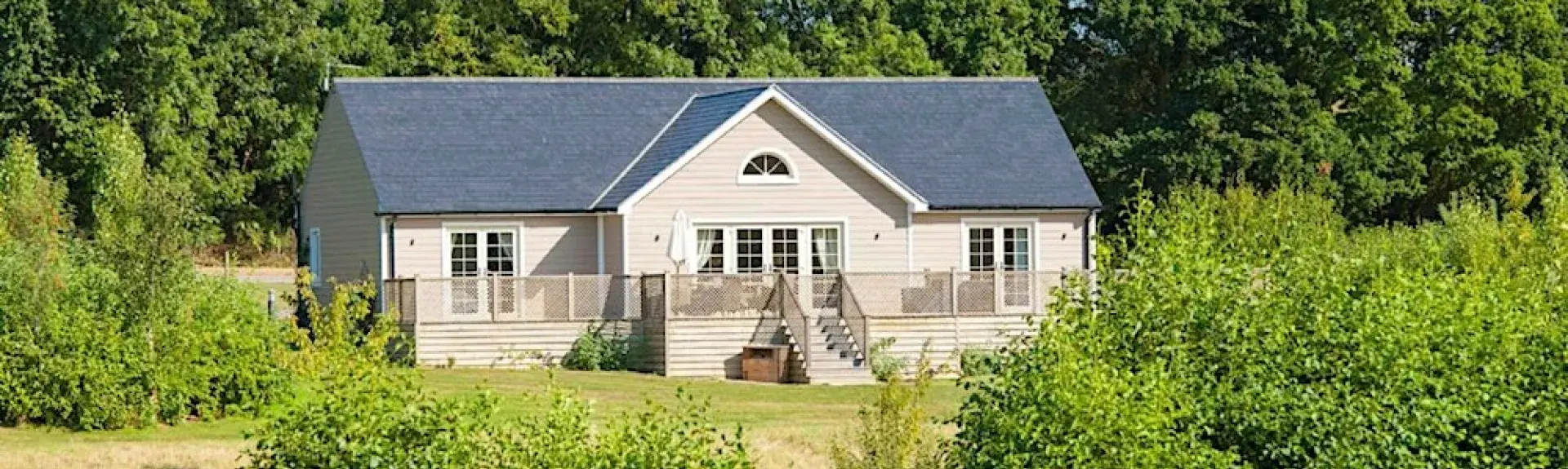 A modern single storey lodge stands in front of trees overlooking a hedge-lined field.