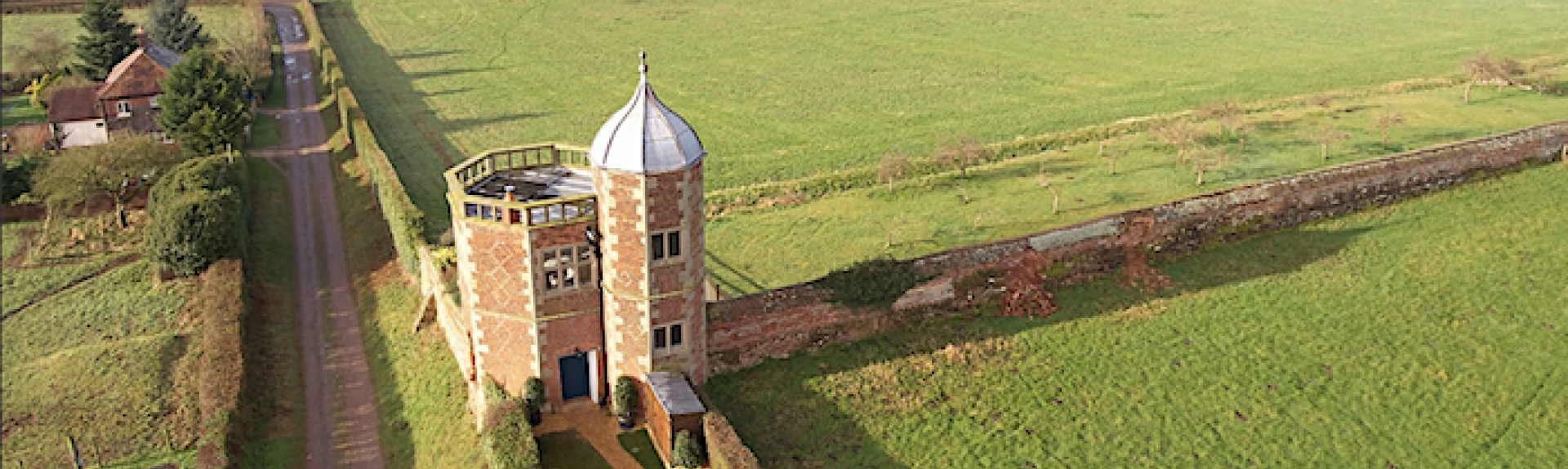 Aerial view of a tall, brick-built, twin-towered, Victorian folly in a rural location.