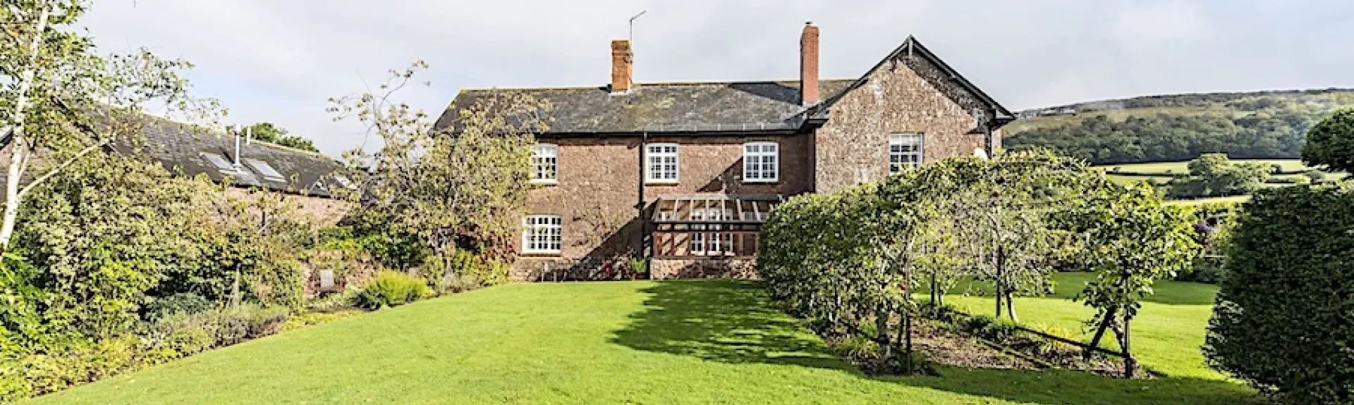 A stone cottage surrounded by fields overlooks a large garden with apple trees.