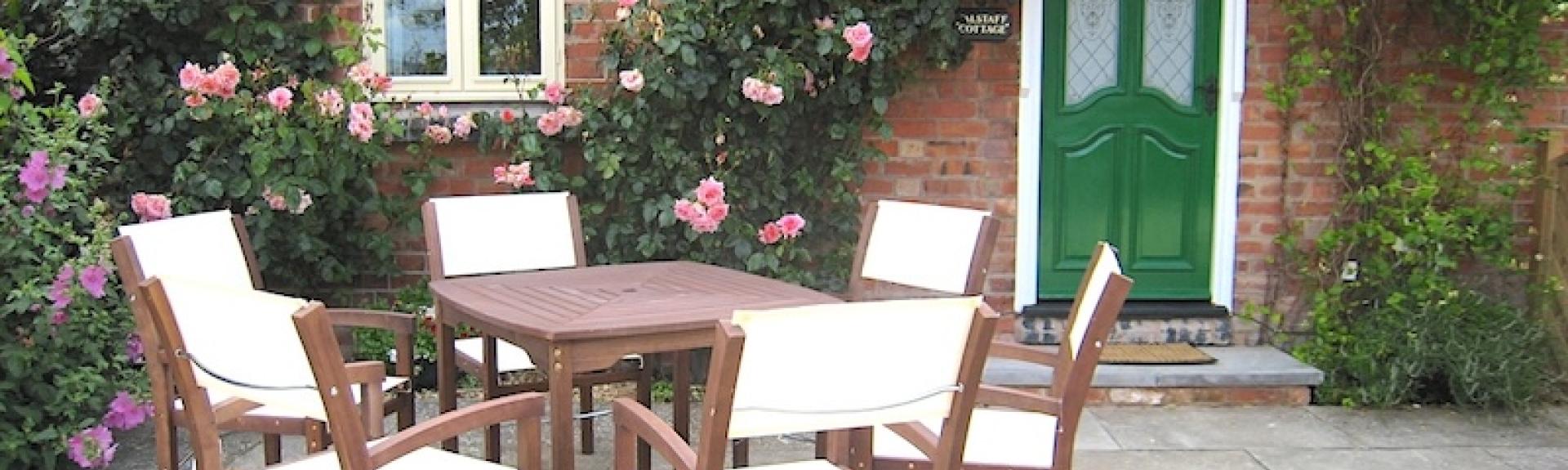 Exterior of  brick-built cottage clad with a rambling rose and overlooking a paved area with dining tsable and chairs