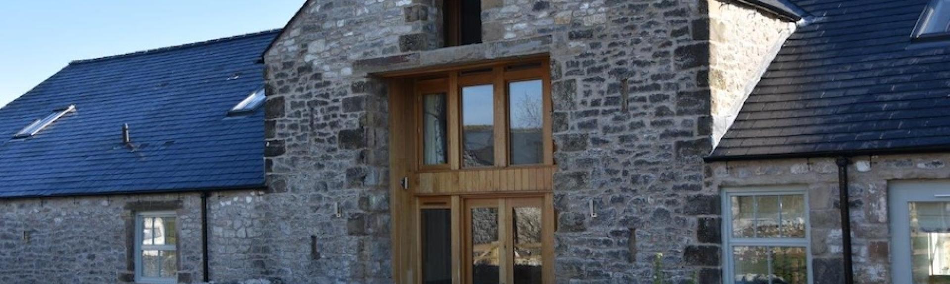 Exterior of a stone-built barn conversion with floor-to-ceiling windows behind a low stone wall.
