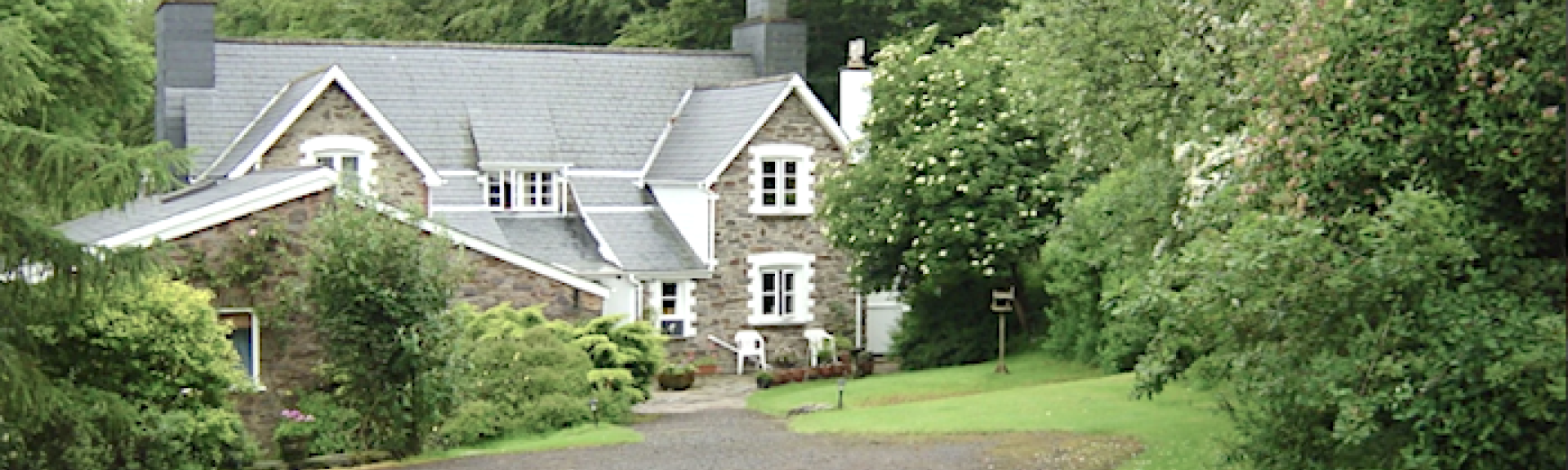 A sweeping drive approaches a twin-baled Exmoor cottage half-surrounded by mature trees.