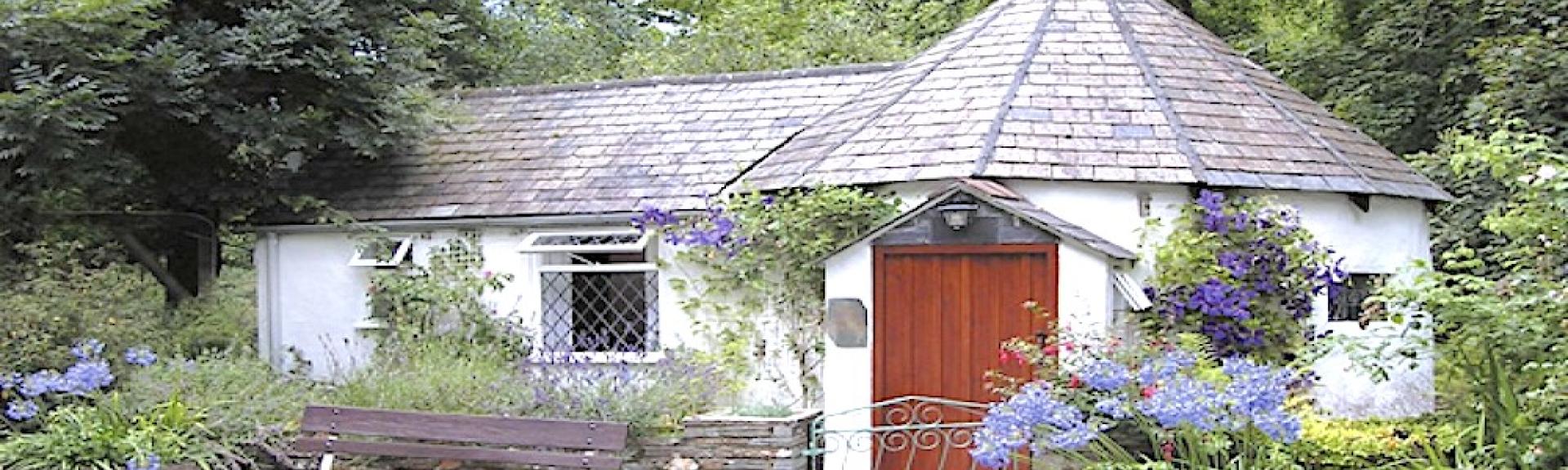 Exterior of a single-storey Cornish cottage surrouded by a tree-lined garden.
