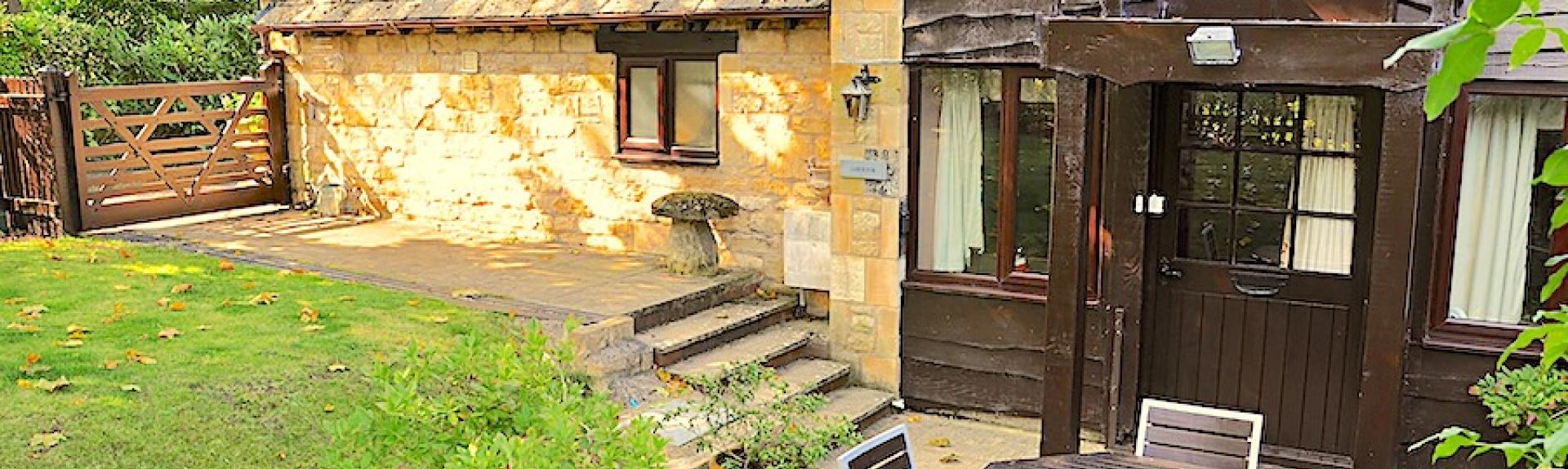 Exterior of a Cotswold honey-stone cottage overlooking a small lawn and patio with a dining table.