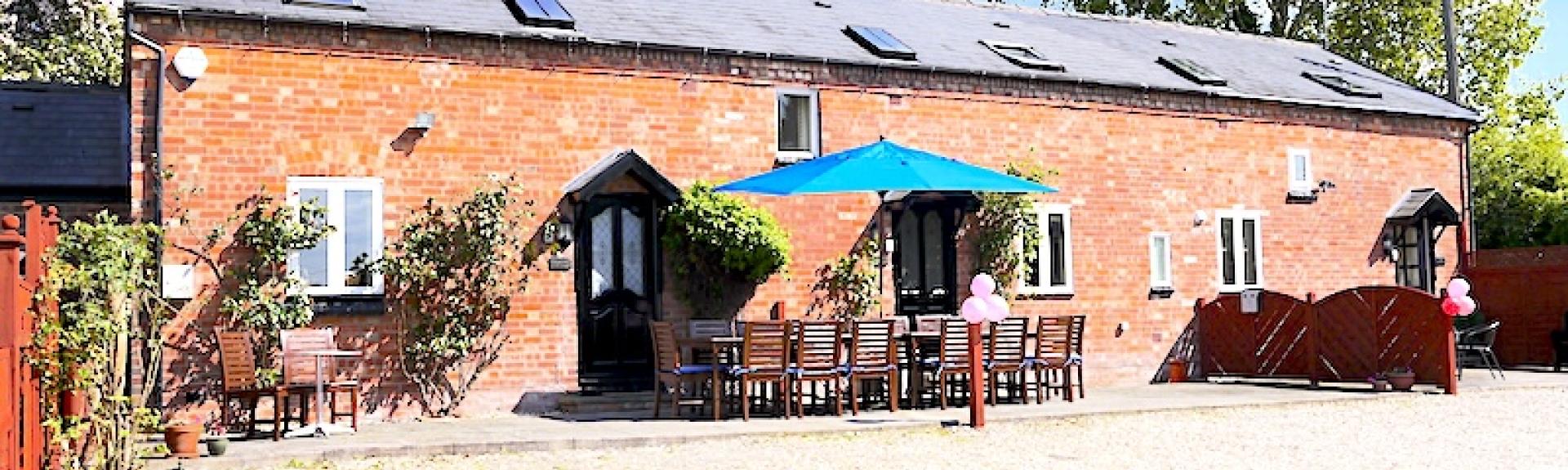 Exterior of a long brick-built barn conversion with a patio with table seating for 14.