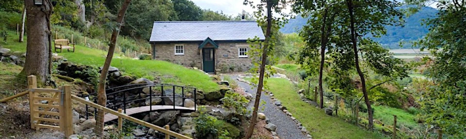 A single storey, stone cottage overlooks a garden with a small stream trickling under a bridge in the foreground.