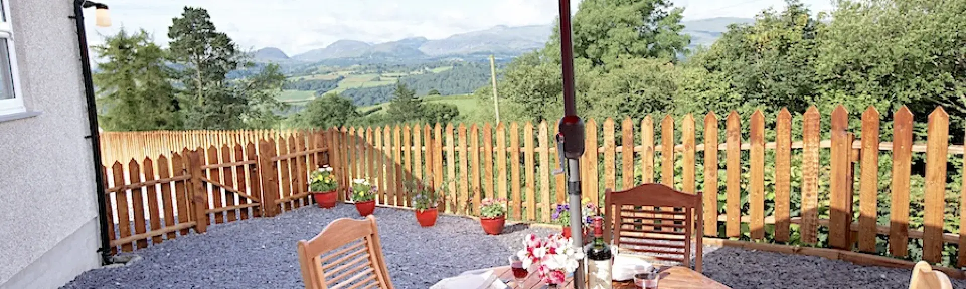 An outdoor dining table underneath a parasol inside a fenced in terrace with rural views beyond. 