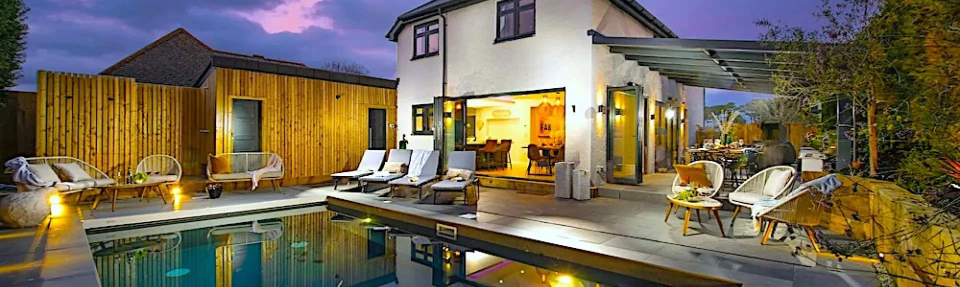 A large, brightly lit house overlooks a swimming pool at dusk.