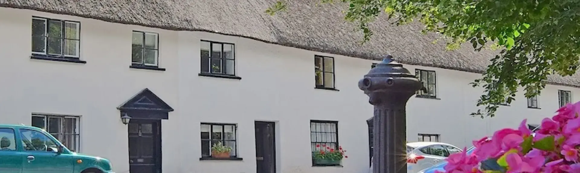 A row of thatched village cottages overlook trees and flowering bushes on a village green.