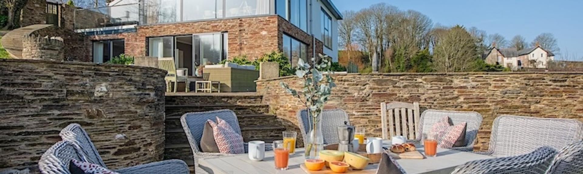 A contemporary country house overlooks a terrace with table laid for meals.