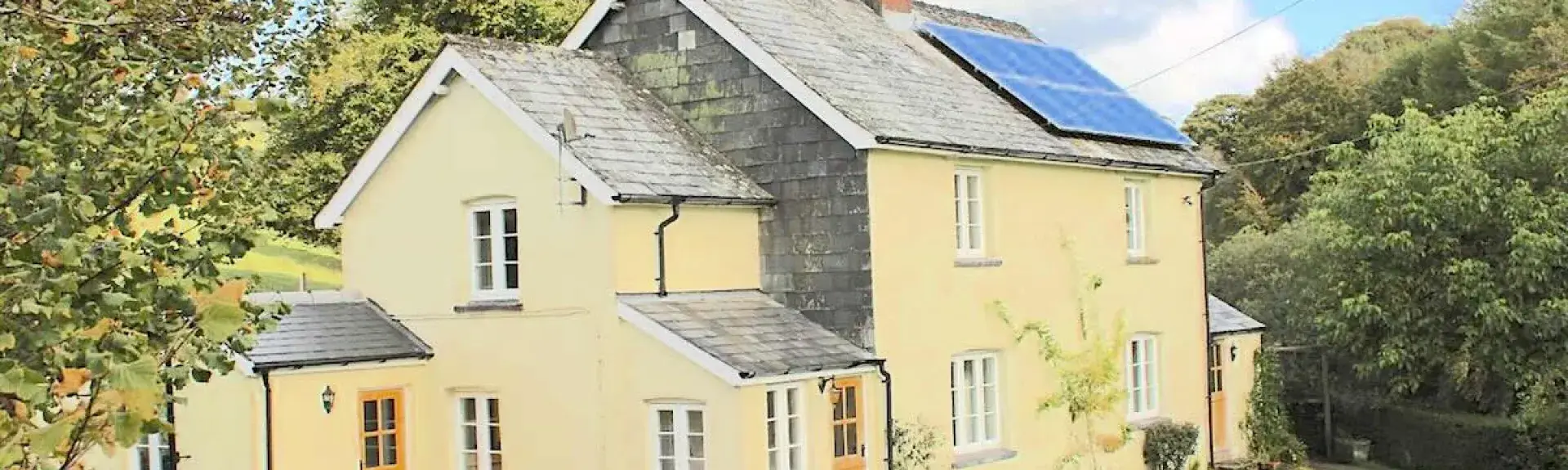 A 2-storey-cottage overlooks a lawn surrounded by trees.