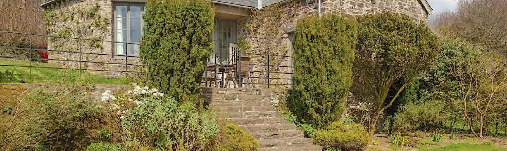 Large single-storey stone-built barn conversion half-hidden by tall shrubs.