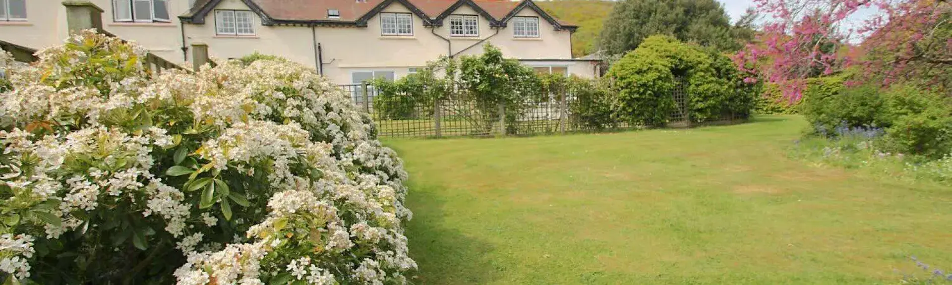 A large Someerset house overlooks a spacious lawn lined by flowering shrubs and trees.