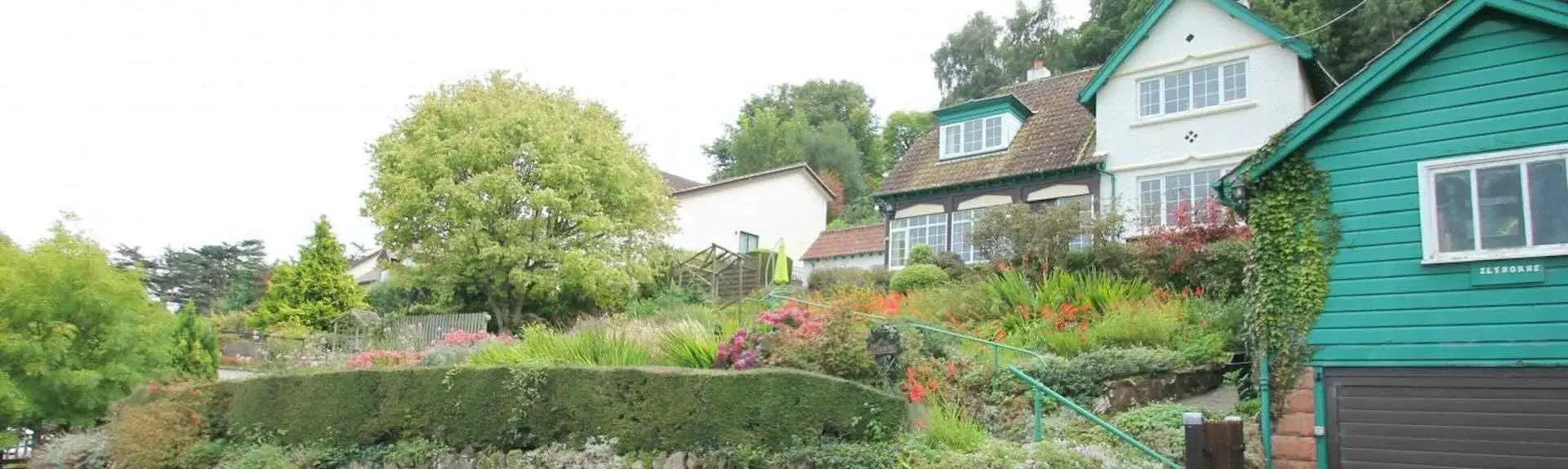 A mature garden slopes up to a large house backed by trees.