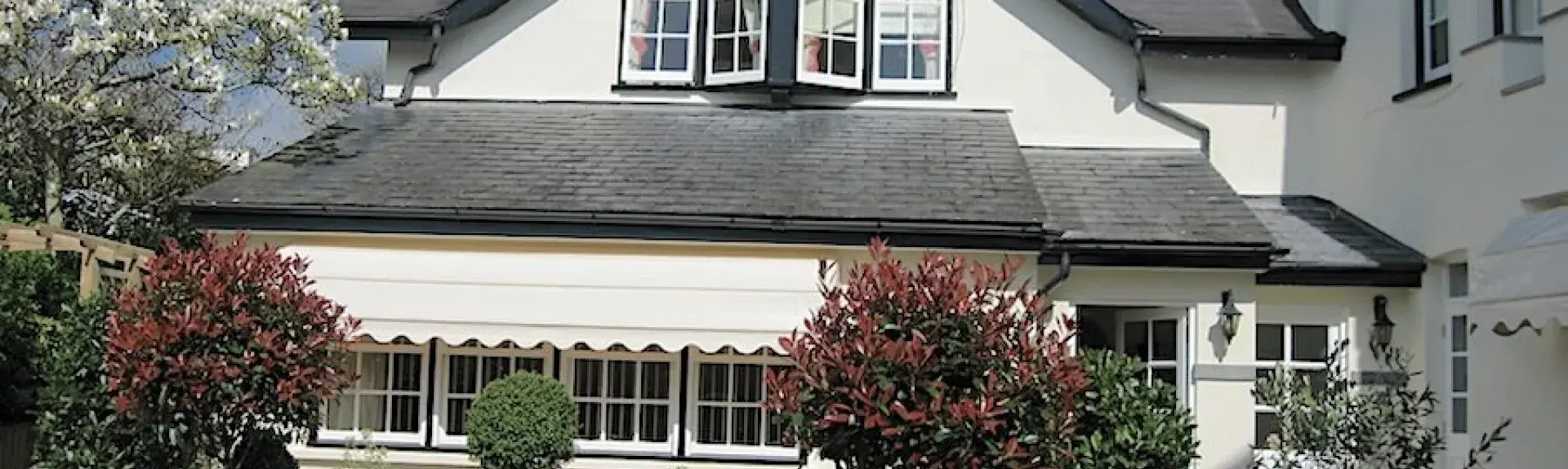 Exterior of a 2-storey cottage with 1st-floor bay windows overlooking a shrub-filled garden.