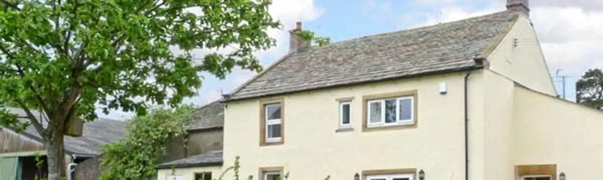 A wood and slate Cotswold holiday cottage overlooks a terrace with a dining table and lawn.