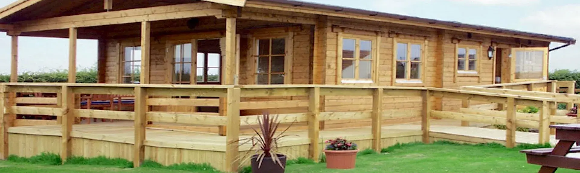 A long eco-friendly log cabin with a covered, wrap-around deck is surrounded by countryside..