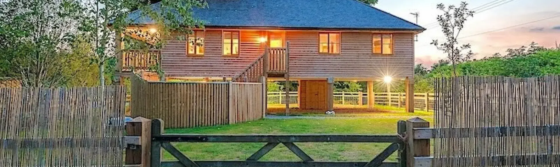 A timber house on stilts beneath lime trees overlooks a large fenced garden.