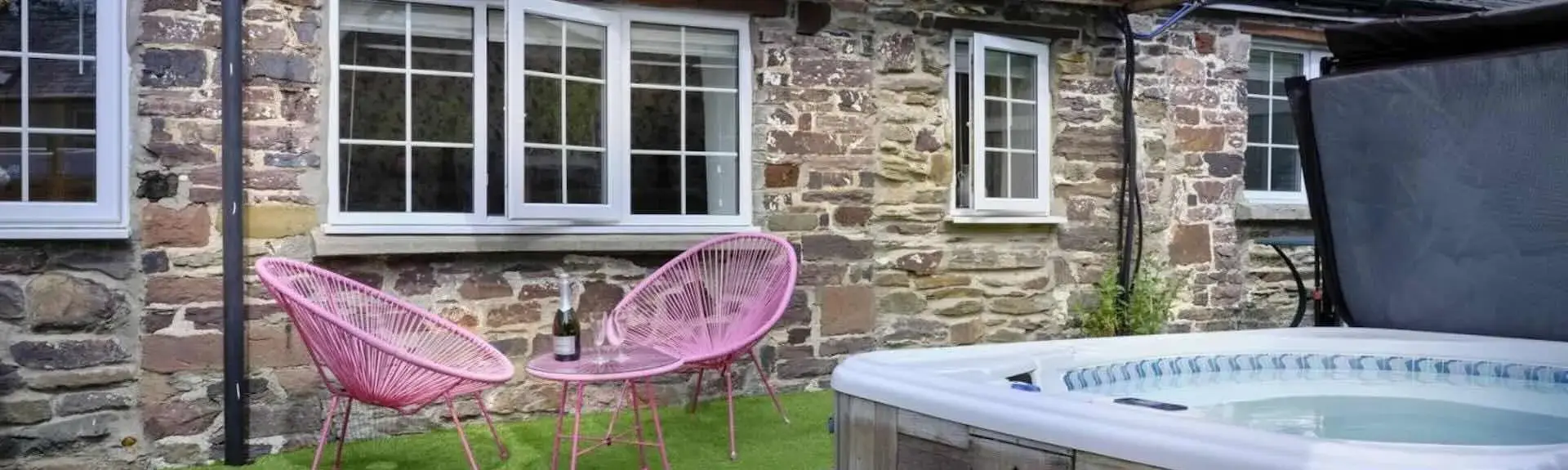 A stone built Exmoor cottage  faces a lawn containing a sheltered hot tub  and a two outdoor chairs around a tea-table.