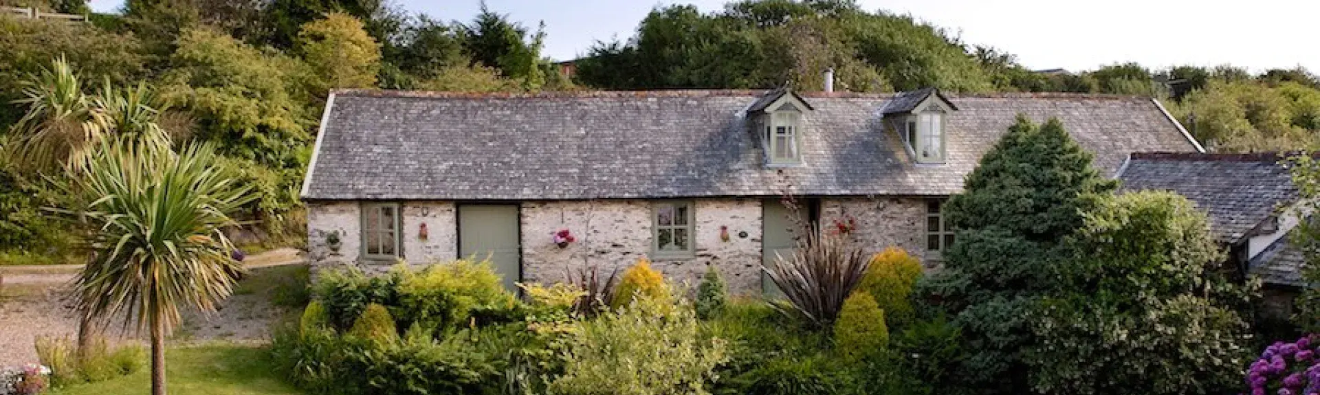 A stone cottage overlooks a lawn bordered by flowering shrubs.