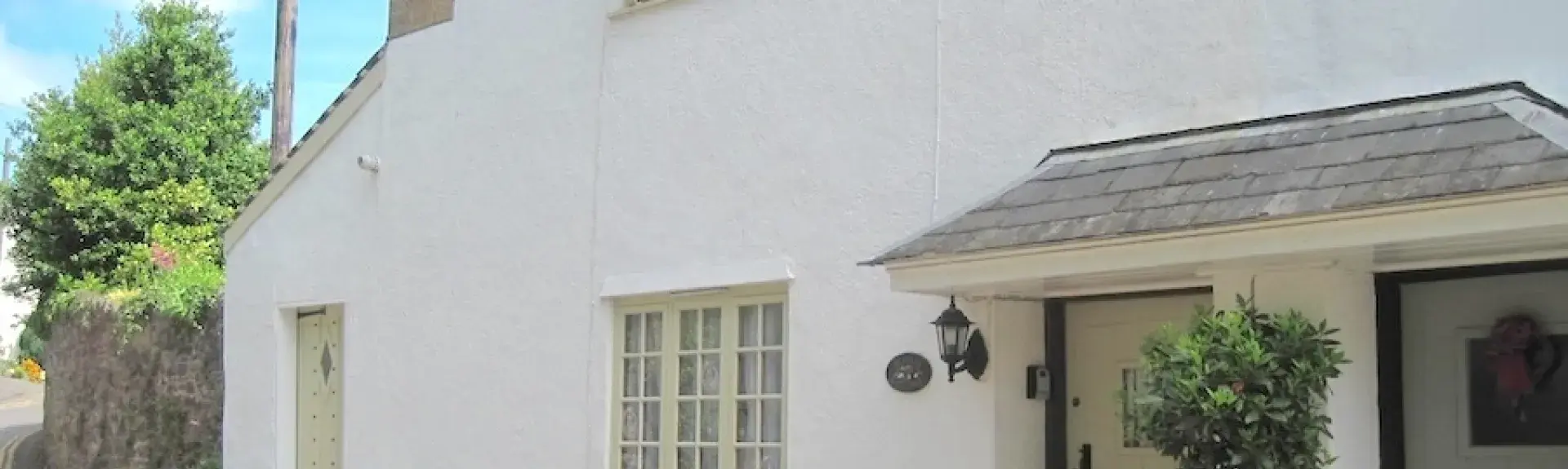 The exterior of a village holiday cottage with a sheltered porch and flower-filled planters on a village street.