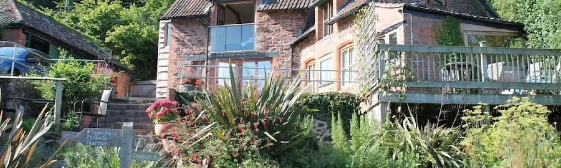 Against a woodland backdrop, a large cottage with a spacious deck overlooks a flower and shrub-filled garden surrounding a lawn.