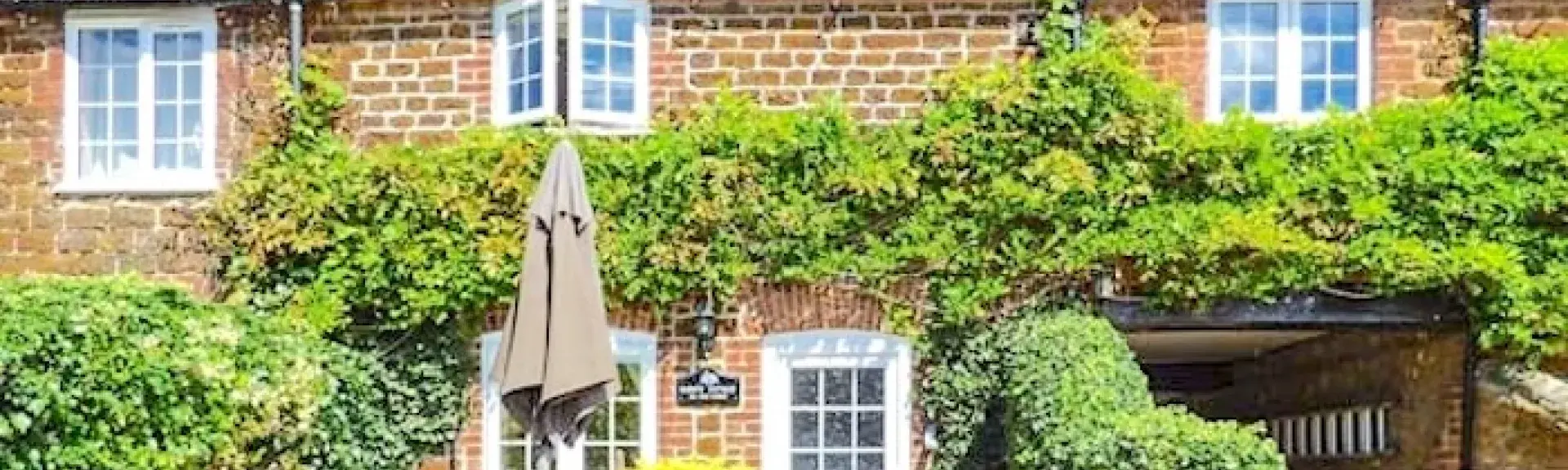 Exterior of a wisteria-clad cottage-front facing a paved front-garden with outdoor dining table, chairs and parasol.