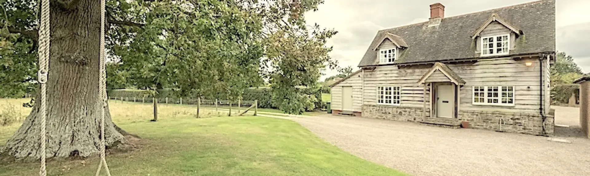 A garden with a garden swing, trees and a wide drive are overlooked by a weather-boarded 2-storey Herefordshire cottage.