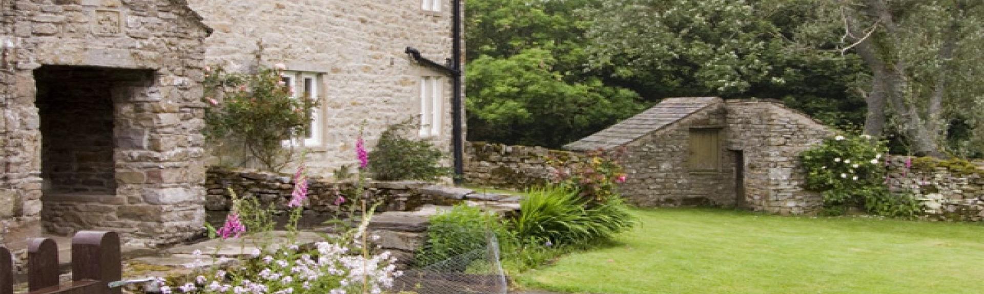 A stone cottage with a large porch overlooks a walled lawn and flower beds.