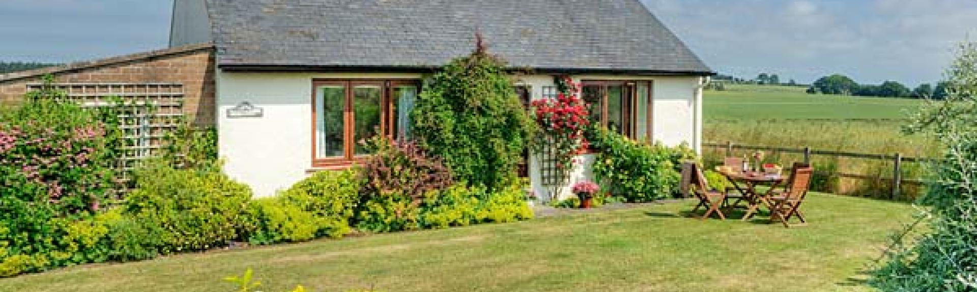 A holiday bungalow with a flowerbeds and a lawn surrounded by open fields.
