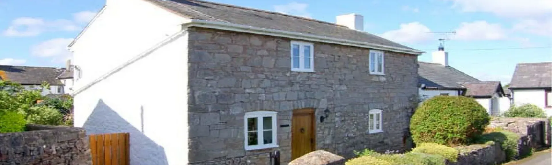 A double-fronted stone cottage  with a hedged-in lawn to the front and a gated driveway for cars.