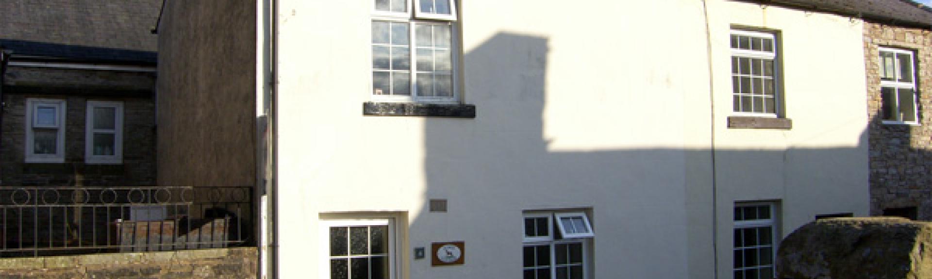 A white-rendered 2-tory, semidetached holiday cottage in Cumbbria overlooks a quiet lane.