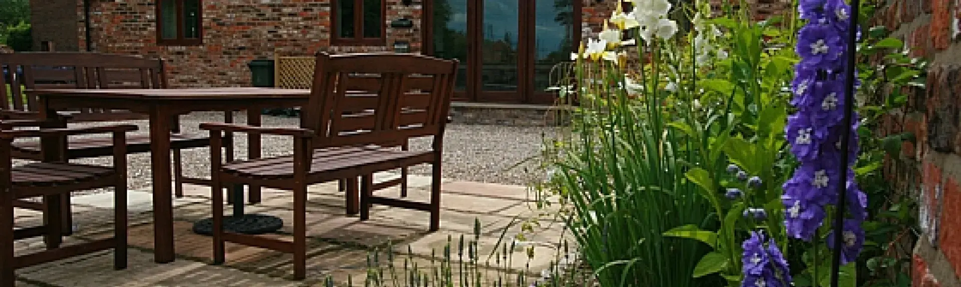 Single-storey barn conversion overlooks a spacious courtyard with dining furniture bordered by flower beds.