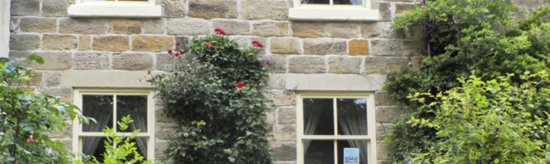 A semi-dtached, stone-buit North Yorkshire holiday cottage sits behind afront garden with shrubs and roses.