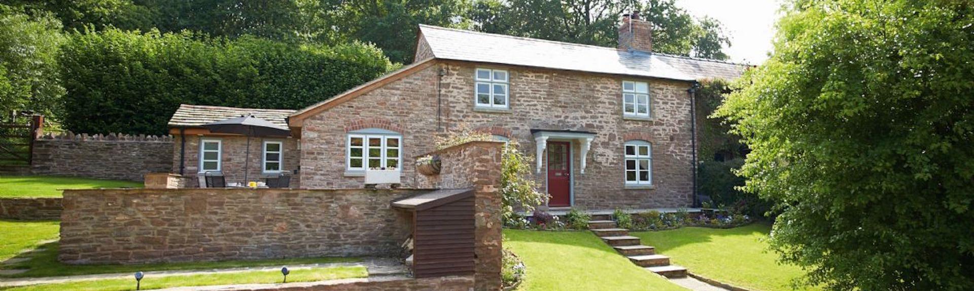 A stone-built Herefordshire cottage overlooks a tree-lined lawn and parking space.