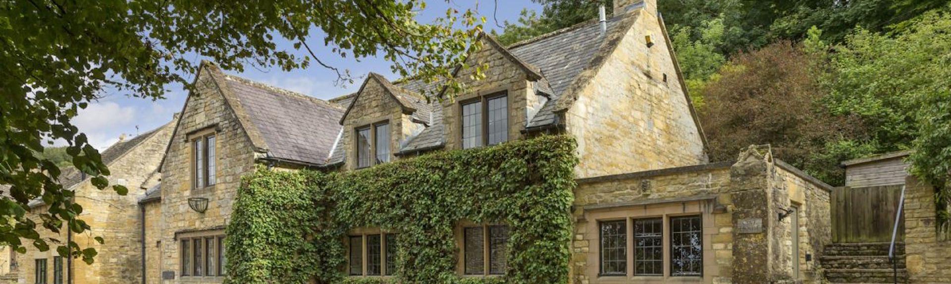 A 2-storey, Cotswold honeystone cottage backed by tall trees.