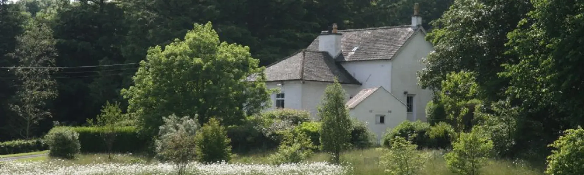 A country cottage on the edge of a wood surrounded by well-kept lawns and flower beds.
